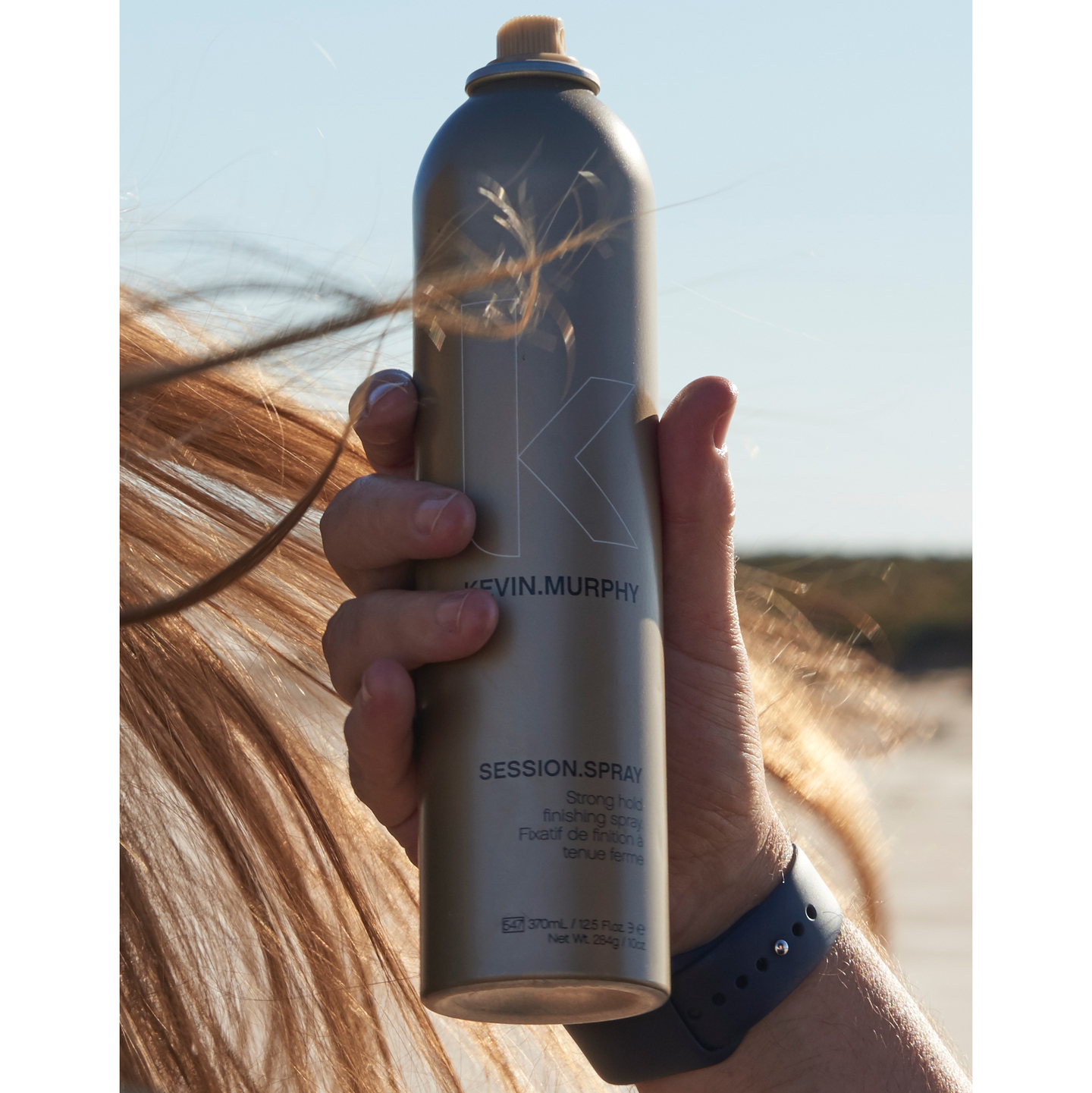 Hand holding a KEVIN.MURPHY SESSION.SPRAY against a windy beach backdrop.